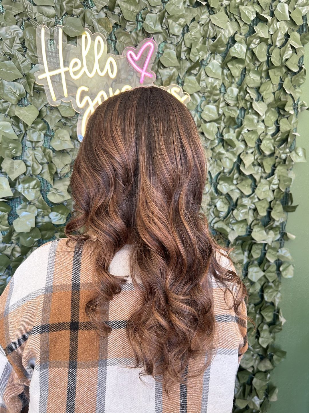 After: warm honey balayage styled in soft waves, in front of the salon's green leaf wall and 'hello gorgeous' neon.